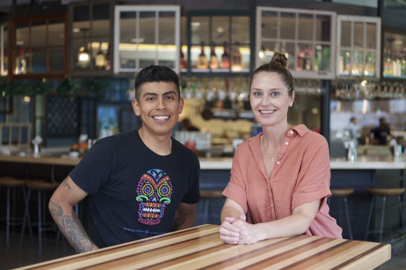 Two people sit at a table with a bar in the background. They are smiling. The one on the left has a black T-shirt and tattoos, the one on the right is wearing a pink button down. 