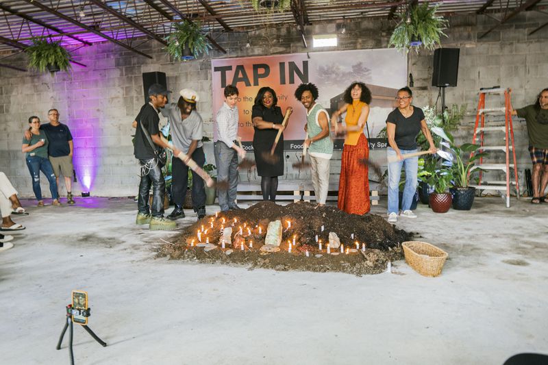 A group of people stand in a cement building and shovel dirt into a mound on the floor that’s lit with candles. 