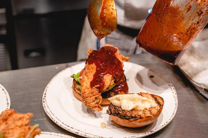 A fried chicken sandwich on a white plate drizzled with rich red sauce.