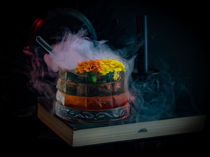 A green, red, and black cauldron garnished with marigolds overflows with dry ice smoke. It is place on top of a book. 