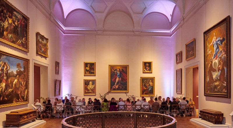 A large room in a museum with high ceilings and white walls. There are paintings on the walls and a long dining table with a group of people seated at it.