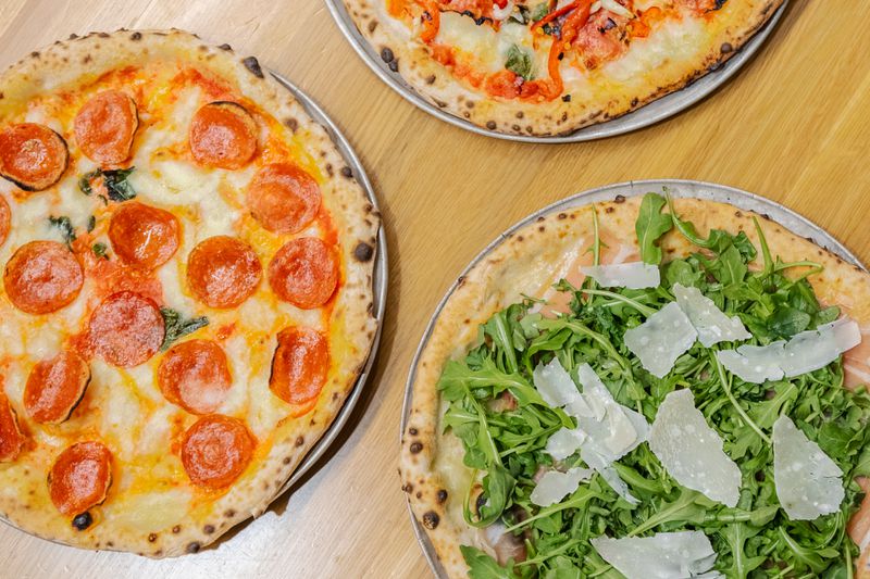 A pepperoni pizza and a pizza topped with arugula and parmesean shavings are on a light wooden table. 
