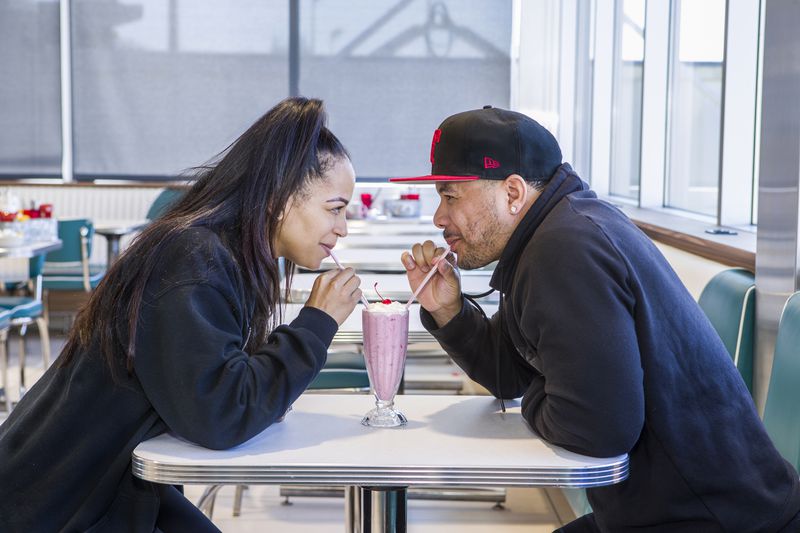 Two people wearing black sweatshirts smile at each other as they both sip from straws in a pink shake with whipped cream and a cherry. 