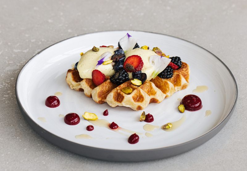 A waffle tipped with berries and flowers on a white plate with dollops of berry sauce. 