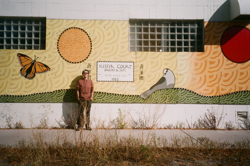 Michelle Kwan stands in front of the colorful Keefer Court mural on the exterior of the store, with has a butterfly and a dove against a yellow and green background. 