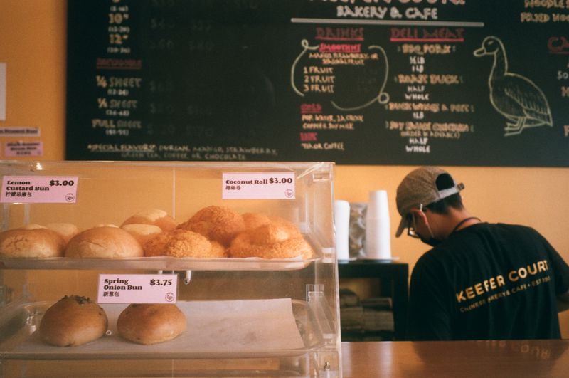 Michelle Kwan stands behind the Keefer Court counter with her back to the camera. In the foreground is a pastry case of coconut rolls, spring onion buns, and lemon custard buns.