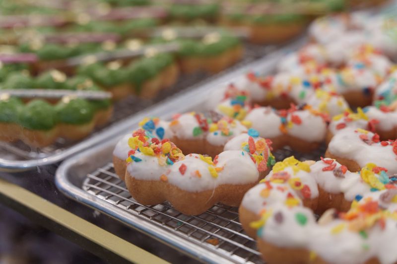 White mochi doughnuts topped with Fruity Pebbles and other mochi doughnuts topped with green frosting in the background. 