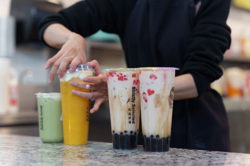 A person dressed in black puts the lid on a tall orange drink. Also on the counter are two tall drinks with boba and red lychee, and a smaller green drink.