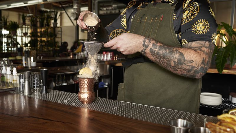 A tattooed bartender dusts powder over a cocktail in a copper class at a bar. 