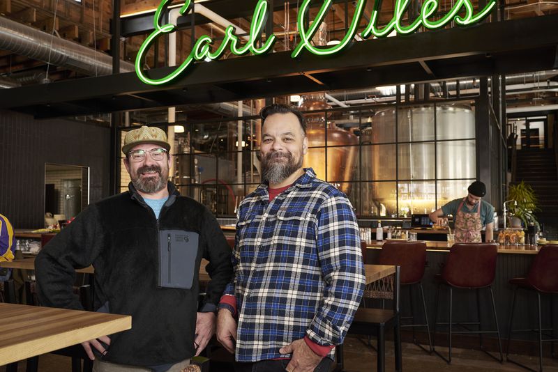 Jeff Erkkila and Jesse Held stand in their distillery, in front of a glowing green Earl Giles sign. 