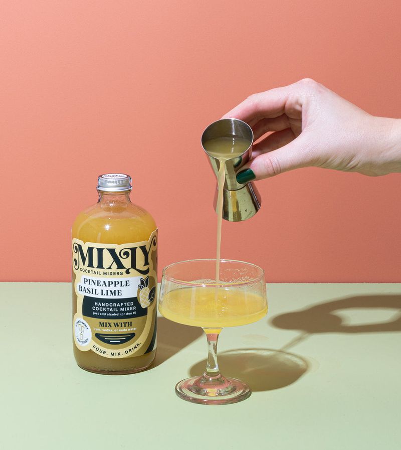 A hand pouring yellow liquid into a cocktail glass, and a bottle labeled “Mixly Pineapple Basil Lime” against and orange and pink background. 