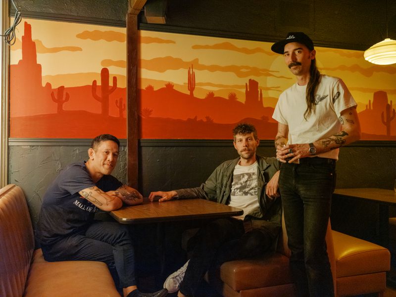 Dan Manosack, Travis Serbus, and Bennett Johnson in a booth at their bar and restaurant, Little Tijuana. There’s a desert motif on the wall. 