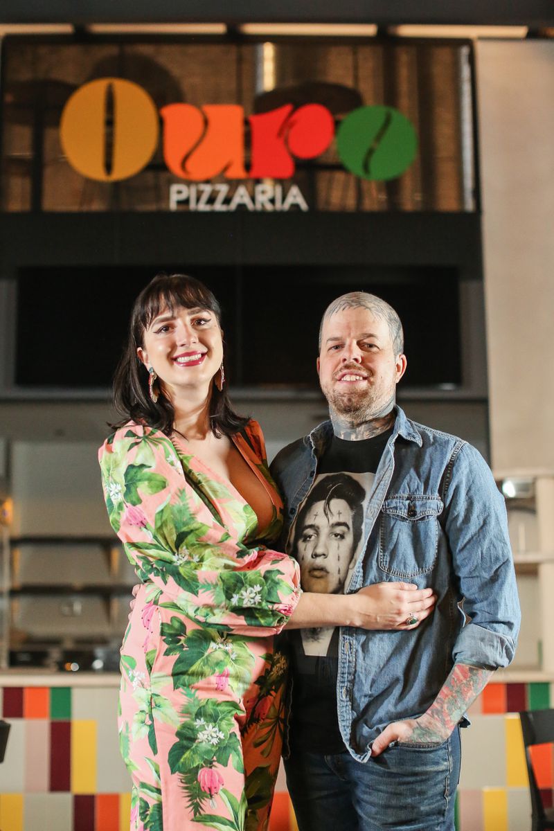 A woman in a green and pink dress and a man in a jean shirt stand in front of a food stall with a large sign reading “Ouro Pizzaria” in shades of orange, red, and green.