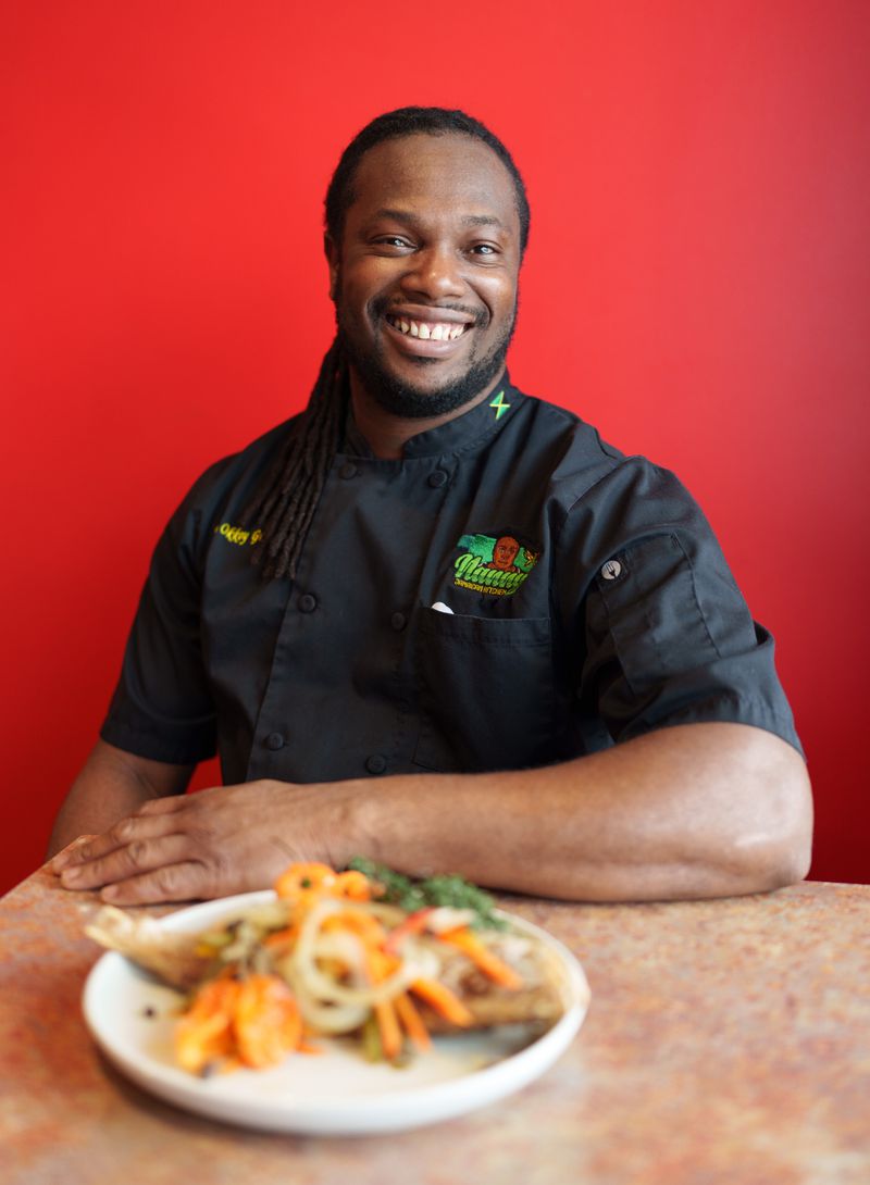 Chef Okkoy Graham in a black chef’s coat sitting in front of a red wall at a table with a dish of escovitch fish in front of him. 