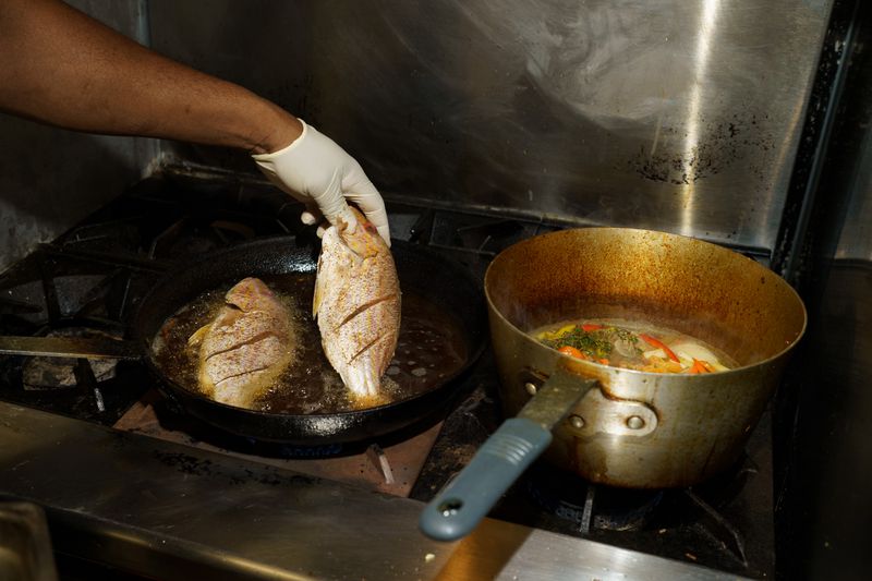 A hand lifting a whole fish out of a frying pan. There’s another fish in the same pan, and next to it, a pot of vegetables stewing. 