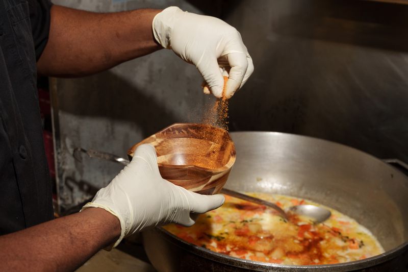 Hands grabbing spices out of a wooden bowl, with a large pot of vegan stew in the background.