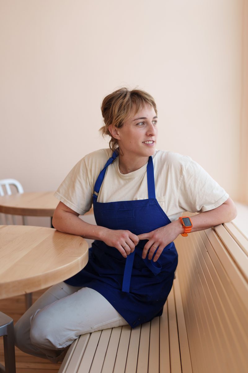 Martha Polacek, wearing a white T-shirt and blue apron, sits on a wooden bench at a table, looking to the right. 