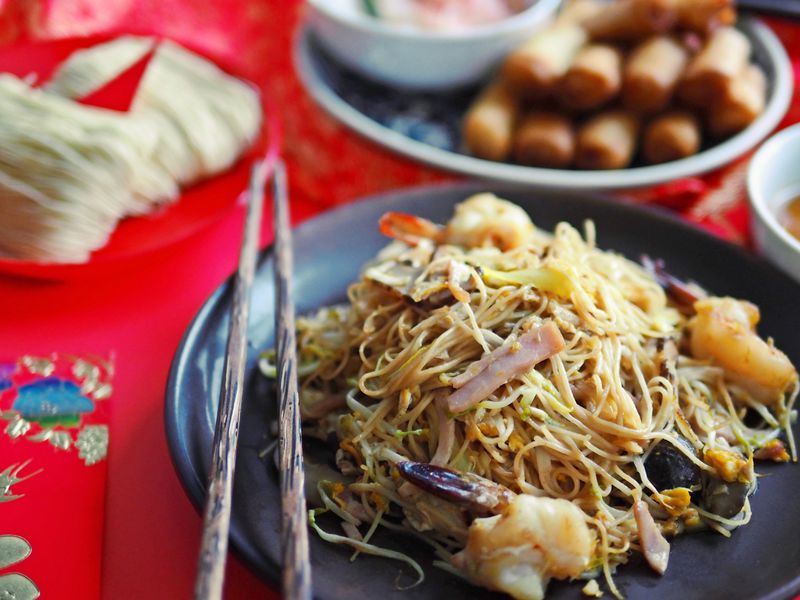 A dish of noodles with meat and shrimp on a red background with egg rolls visible in the background. 