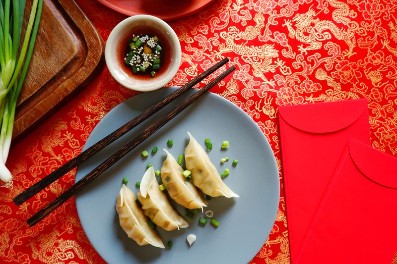 Four dumplings on a blue plate with chopsticks and a small bowl of sauce on a bright red background. 