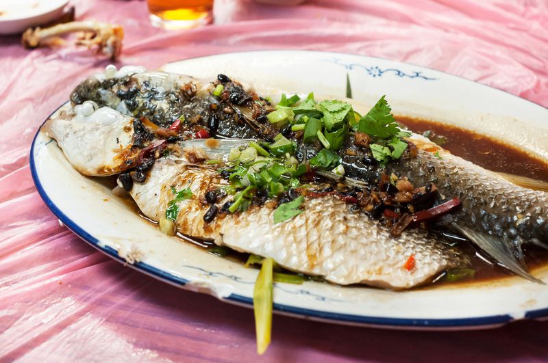 A plate of steamed whole fish in black bean sauce on a pink plastic tablecloth.  