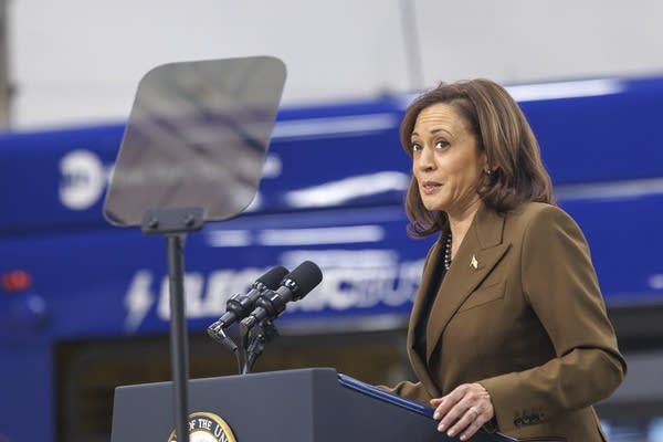 A woman in a brown suit at a podium looks to her left