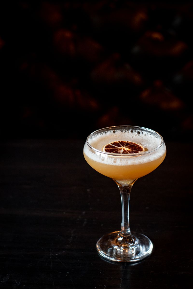 A foamy bourbon cocktail in a gimlet glass topped with a slice of dried orange against a black background. 