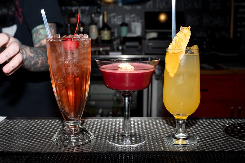 Three cocktails sitting next to one another on a bar. The far left is an amber color, the middle is a deep red, and the far right is yellow with a wedge of dried pineapple.