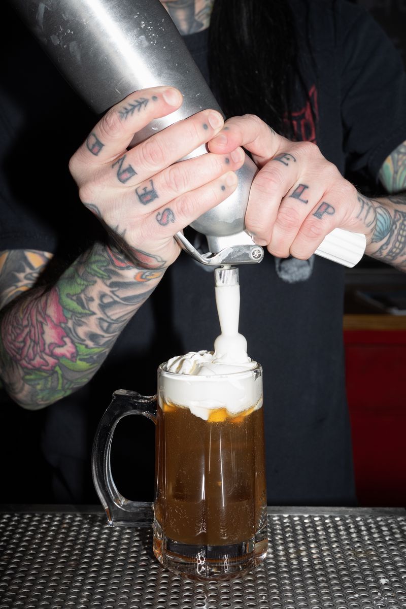 A person squeezing white foam on top of an amber colored cocktail in a rootbeer mug. 