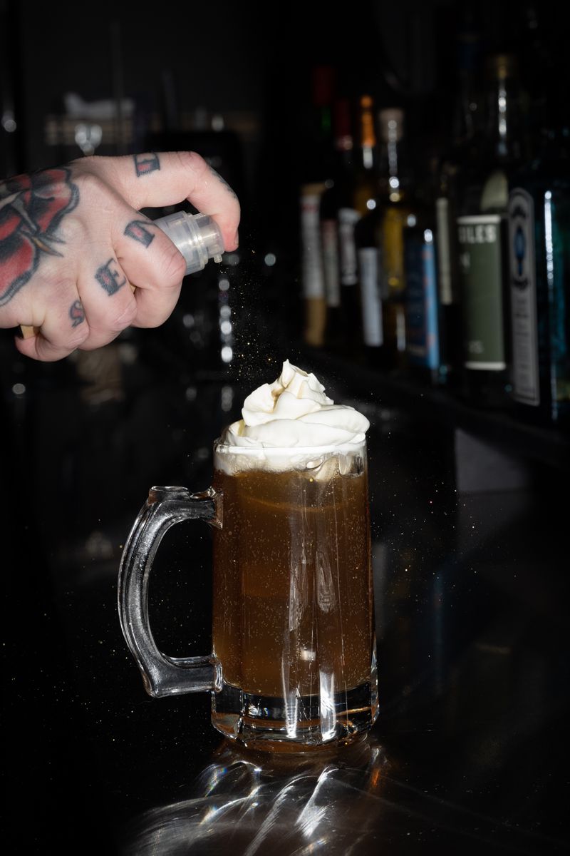 A hand spritzing an amber cocktail topped with white foam in a rootbeer mug glass. 