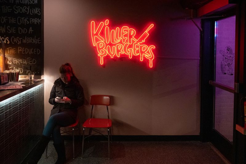 A white wall with a neon sign that reads “Killer Burgers” and a person sitting on a chair underneath it. 
