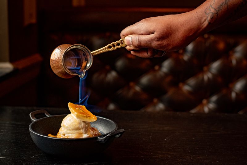 A hand pouring a pitcher of liquid flame onto a bananas foster dish in a small cast iron. 