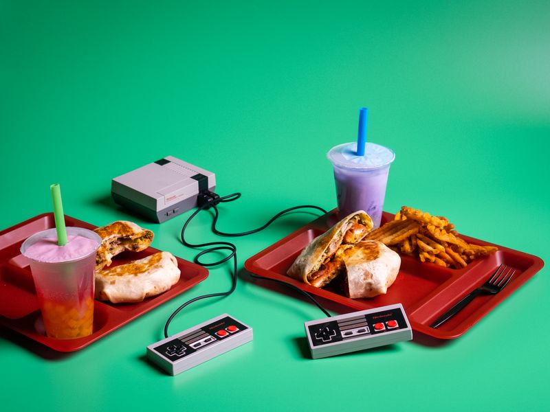 Red school lunch trays with cups of orange and purple bubble tea, tortilla-wrapped pickets, and waffles fries next to a Nintendo video game set, all on a green background. 