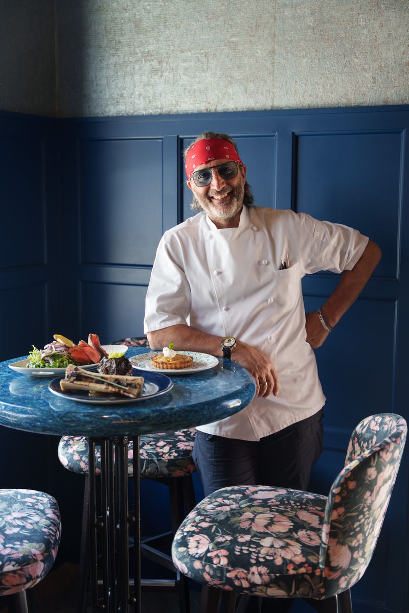 Chef David Fhima wearing a white chef’s coat, a red bandana, and tinted glasses, standing with one elbow on a table with plates of food against a blue wall.