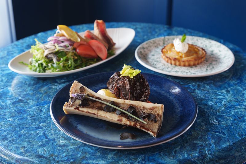 Three dishes on a blue table: A bone marrow beef dish (center), a Nicoise salad (left) and a pear tart (right). 