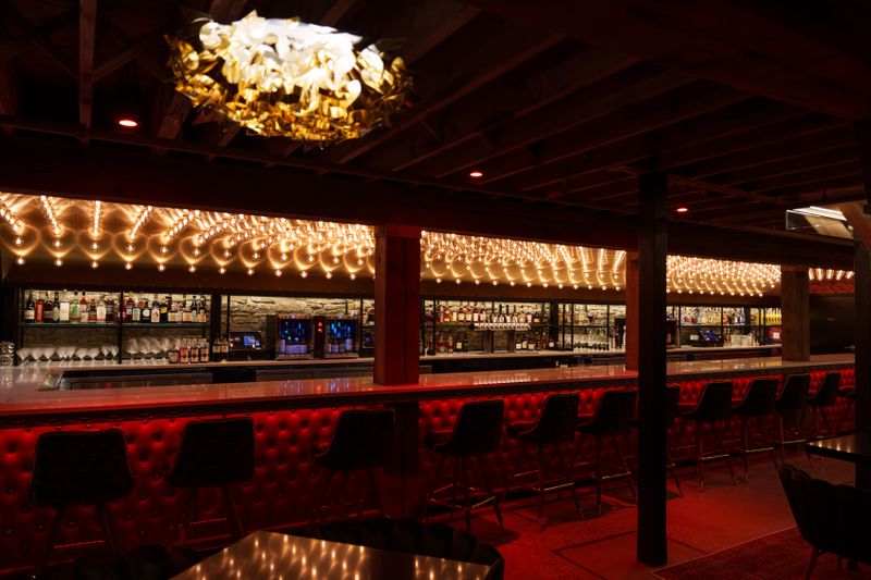 A long bar with marquee lighting on the ceiling, plush red velvet underneath the bar, and a long line of chairs. 