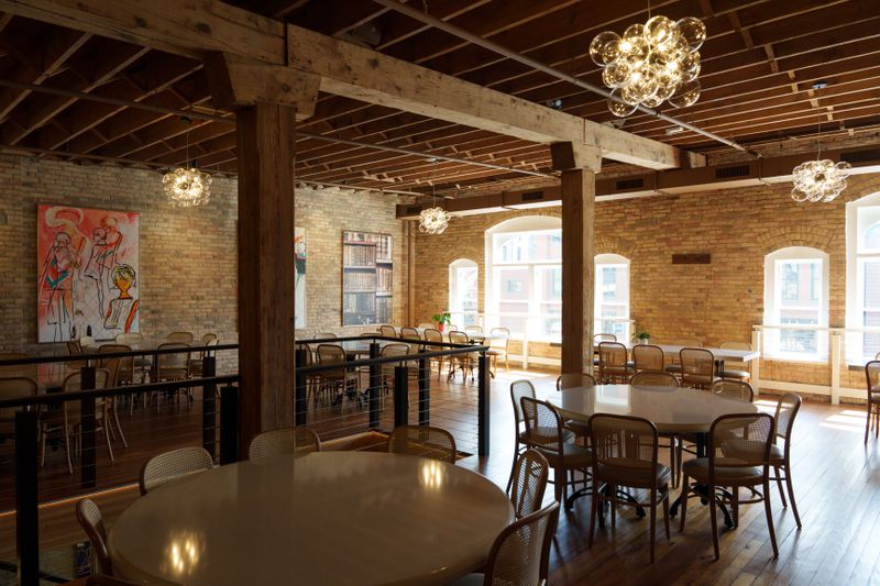 A large sunny room with round tables surrounded by chairs, brick walls, a chandelier, and art on the walls.