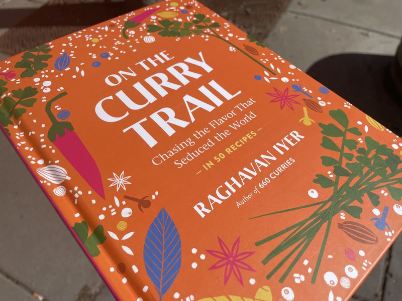 An orange hardcover cookbook with illustrations of food and spices around the border, and this text in white: On the Curry Trail, Chasing the Flavor that Seduced the World in 50 Recipes, Raghavan Iyer, Author of 660 Curries