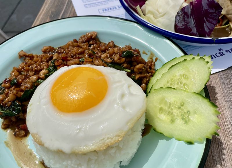Ground chicken and basil leaves next to a mound of white rice topped with an egg with a side of sliced cucumbers on a light blue plate. 