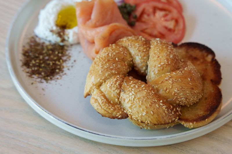 A braided bagel coated in sesame seeds on a plate with za’atar, lox, tomatoes, and labneh. 