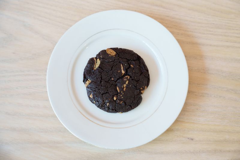 A dark chocolate cookie with caramel chunks on a round white plate on a light wooden table. 