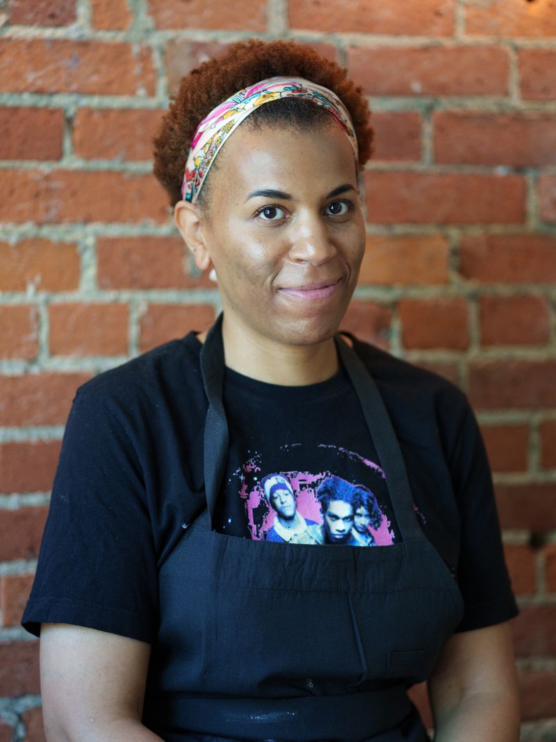Shawn McKenzie wearing a headband, a black graphic T-shirt, and black apron, smiling in front of a brick wall. 