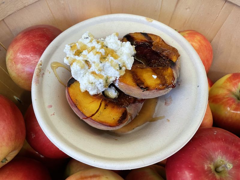 A grilled peach topped with a dollop of goat cheese and honey in a paper bowl sitting in a basket of apples. 