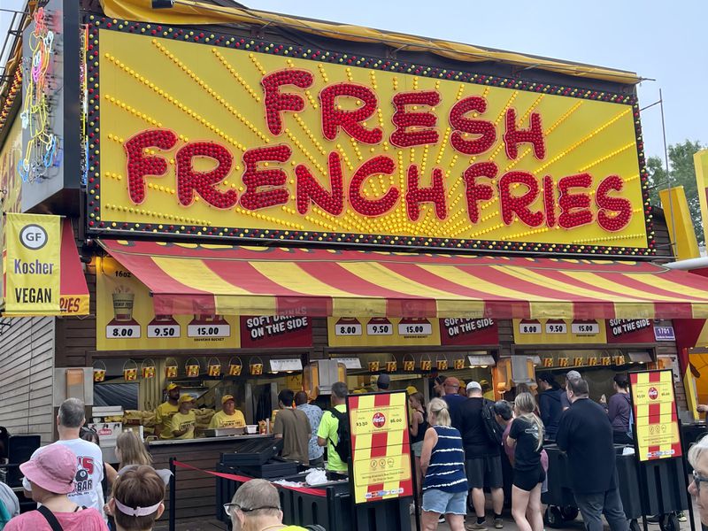 A building with a large yellow and red sign that reads “Fresh French Fries” and a yellow and red striped awning with people standing beneath it. 