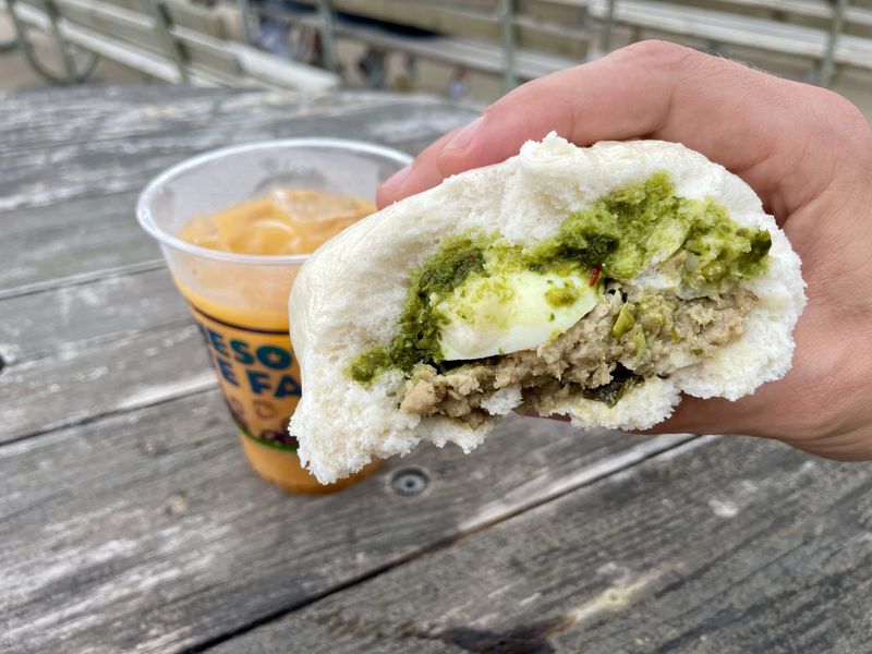 A hand holding a steamed bun stuffed with ground meat, an egg, and green sauce, in front of a cup of Thai tea on a wooden table. 