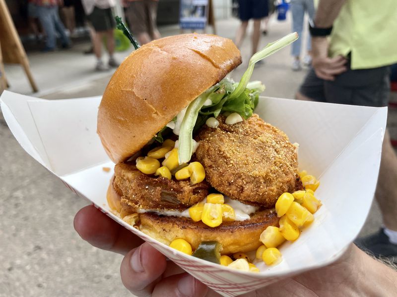 A fried green tomato sandwich topped with corn relish on a golden bun in a white paper dish. 