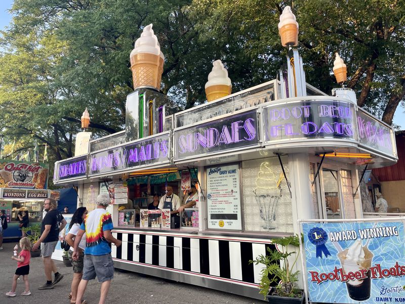 A food stand with purple lettering reading “Malts, Kones, Sundaes” and people standing outside. 
