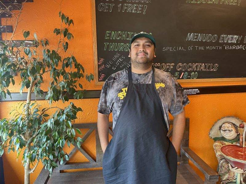 Miguel Hernandez in a black apron wearing a baseball hat in front of an orange wall and chalkboard.