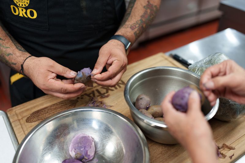 The hands of two people peeling purple potatoes above silver bowls.