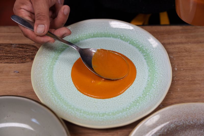 A hand holding a spoon spreading a pool of orange salsa de venas on a light green plate. 