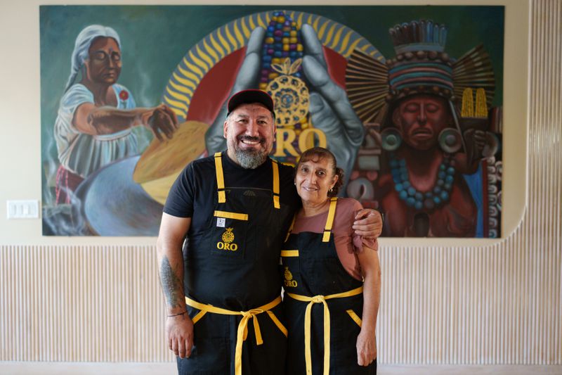 Gustavo Romero and Teresa Veytia Avila wearing black aprons with yellow ties, standing with their arms around each other and smiling. 